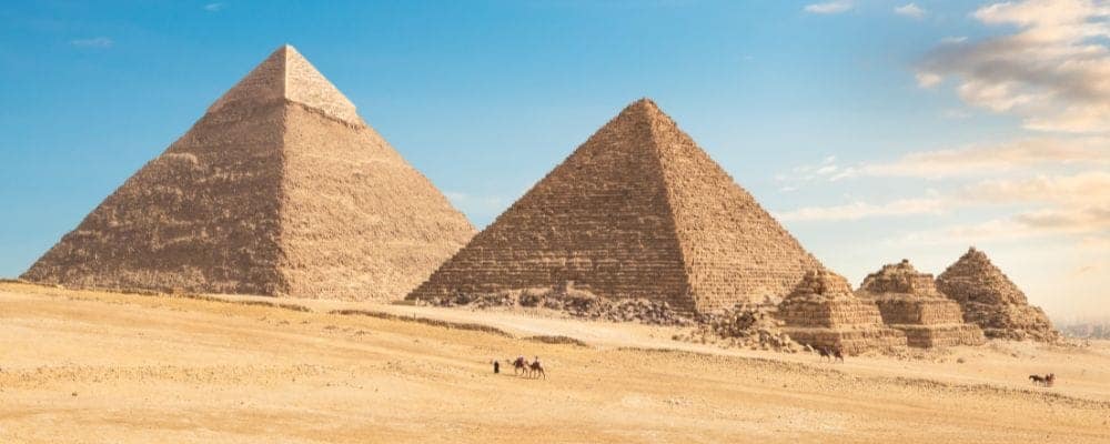 A wide panoramic view of the Great Pyramids of Giza and the Queens' Pyramids with camels in the desert, part of a private luxury itinerary by Top Class Egypt Travel.