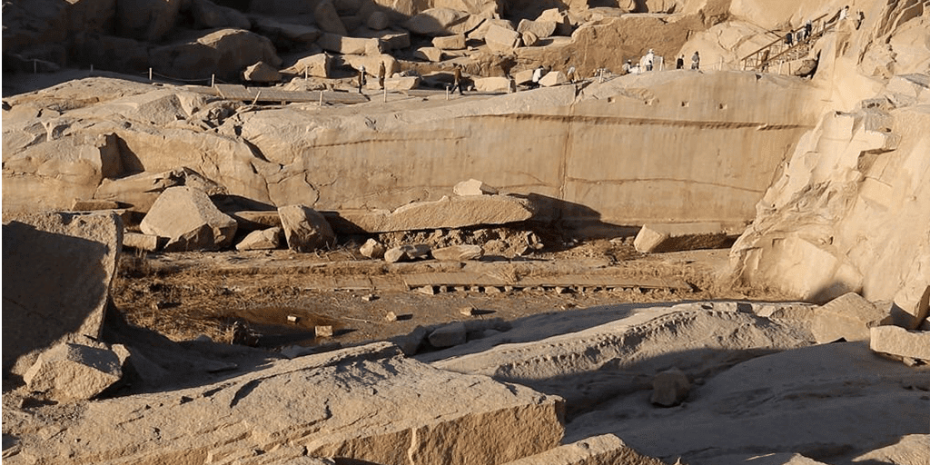 A high-angle view looking down at the massive Unfinished Obelisk carved directly into the pink granite bedrock of an ancient quarry in Aswan, under a clear sky. Caption A fascinating glimpse into ancient craftsmanship: the monumental Unfinished Obelisk