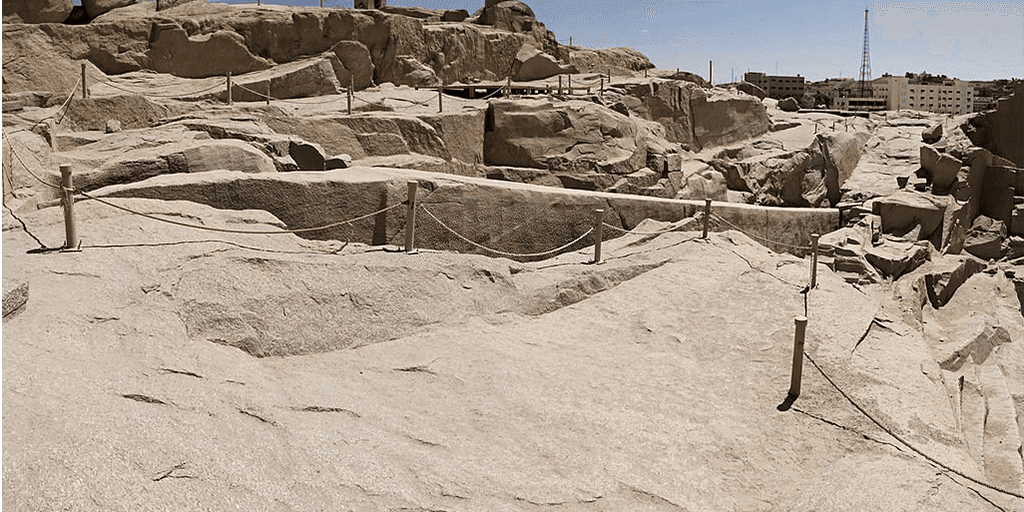 A high-angle view looking down at the massive Unfinished Obelisk carved directly into the pink granite bedrock of an ancient quarry in Aswan, under a clear sky. Caption A fascinating glimpse into ancient craftsmanship: the monumental Unfinished Obelisk