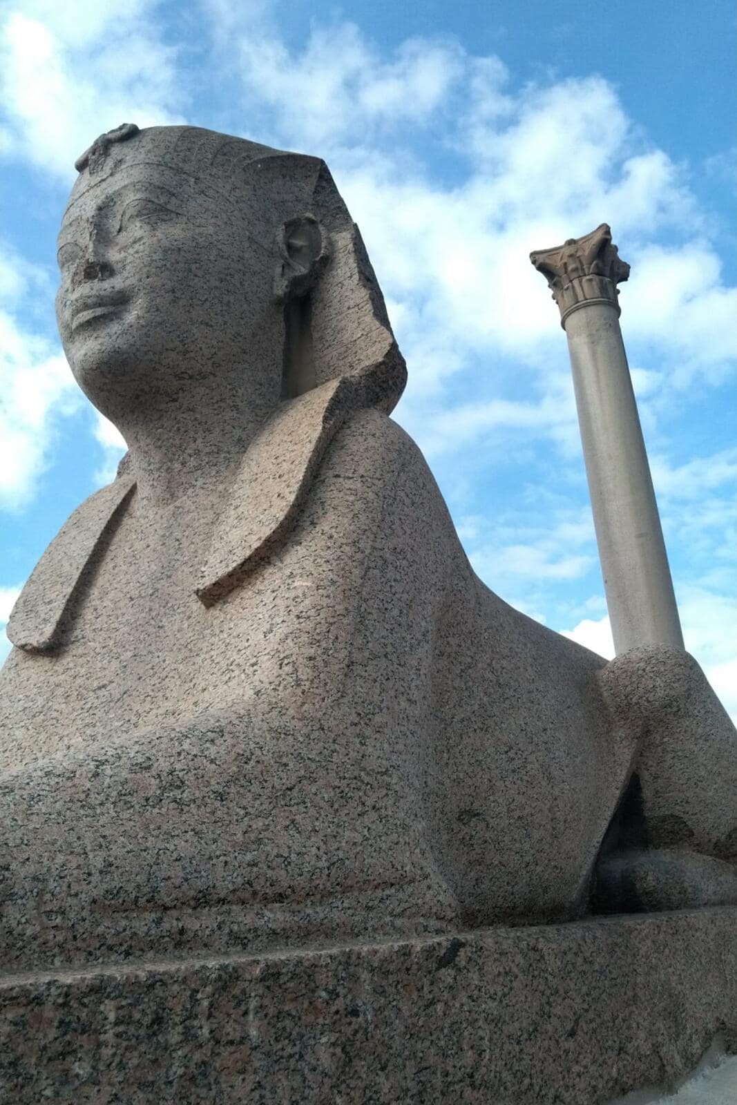 The ancient Roman Pompey's Pillar alongside a granite Sphinx statue under a blue sky in Alexandria Egypt.
