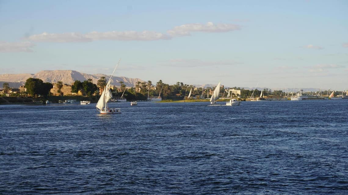 Traditional Egyptian felucca sailboats sailing on the Nile River in Luxor with lush riverbanks and desert hills in the background.9 Days Cairo and Nile cruise.