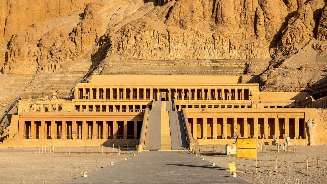 A straight daytime shot of the terraced Mortuary Temple of Hatshepsut at Deir el-Bahari in Luxor, showcasing its grand colonnades and long central ramp built into massive limestone cliffs.