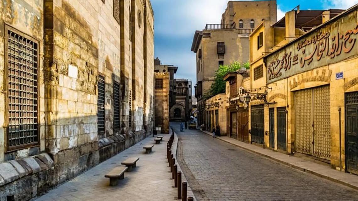 A daytime perspective of the historic cobblestone Al-Muizz Street in Old Cairo, lined with monumental medieval Islamic stone walls, traditional wooden windows, and local shopfronts under a soft sky.