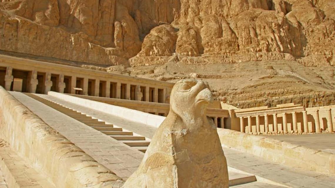 A close-up shot of a weathered stone Horus falcon statue standing guard at the base of a grand stone ramp, leading up to the tiered terraces and colonnades of the Mortuary Temple of Hatshepsut against the steep limestone cliffs of Deir el-Bahari.
