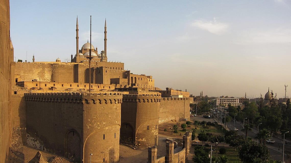 Panoramic view of the historic stone walls of the Citadel of Salah El-Din in Cairo, featuring the prominent domes and minarets of the Mohamed Ali Mosque against a clear sky.