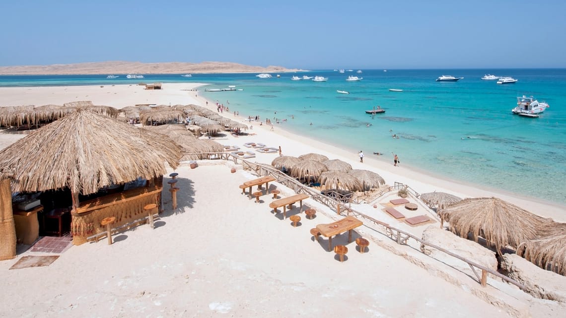A high-angle daytime view of a white sand beach resort in Hurghada, featuring thatched-roof umbrellas, sun loungers, and clear turquoise Red Sea water with boats anchored in the distance.