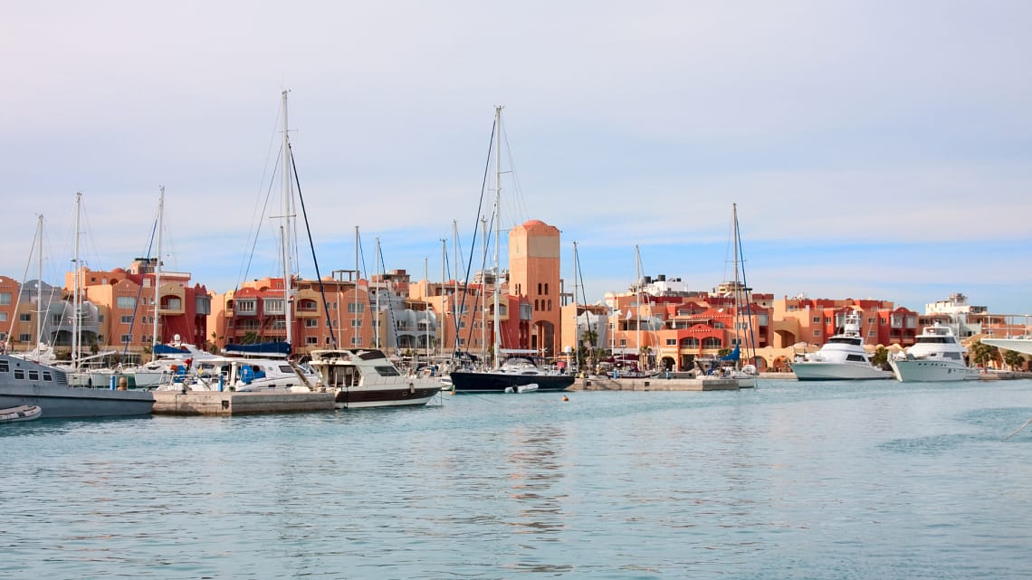 A scenic daytime view of El Gouna Marina featuring luxury yachts and sailboats moored in the calm harbor waters, with Mediterranean-style terracotta and pastel resort buildings lining the waterfront.