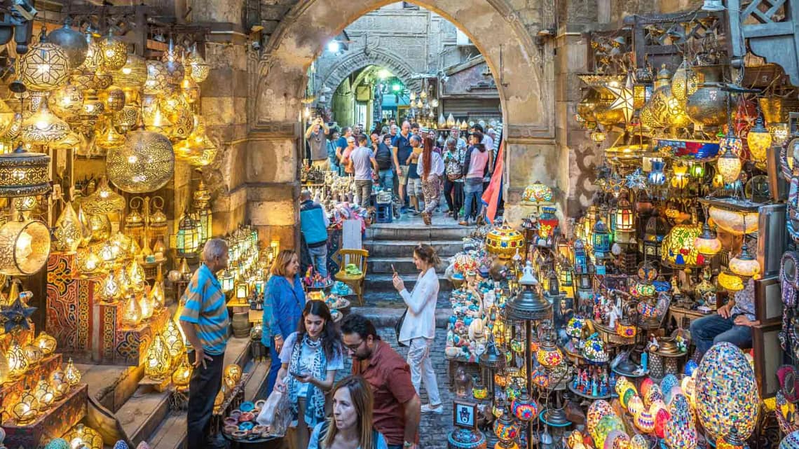 A view through a historic stone archway into a bustling alley of Khan El-Khalili bazaar in Cairo, filled with glittering traditional lanterns, brassware, and colorful artisanal crafts.