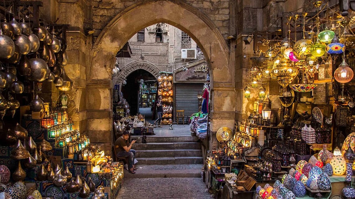 A view through a historic stone archway into a bustling alley of Khan El-Khalili bazaar in Cairo, filled with glittering traditional lanterns, brassware, and colorful artisanal crafts.