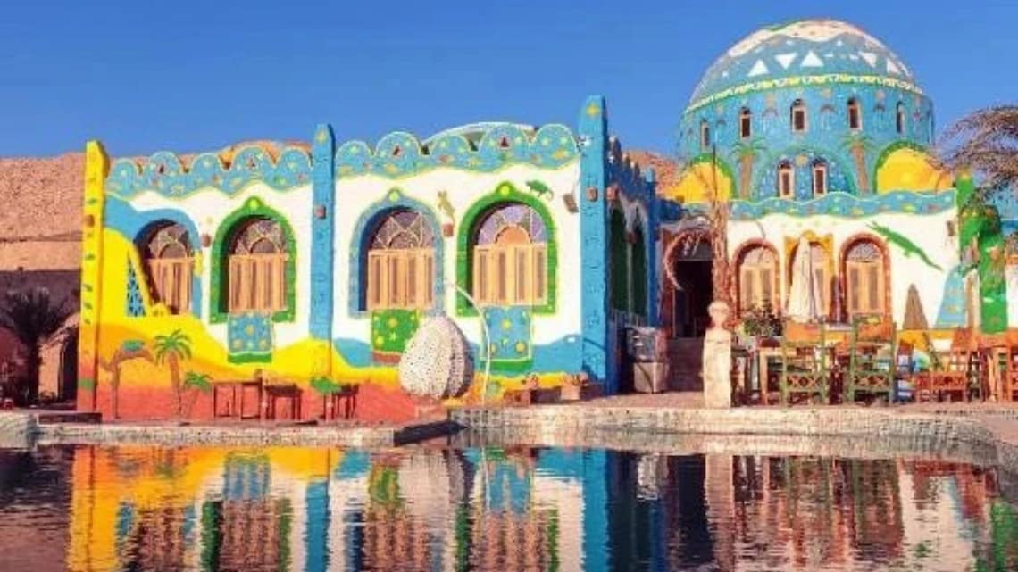 A view from the Nile River of a vibrant Nubian village in Aswan, showcasing traditional brightly painted blue and white houses along the rocky shoreline.