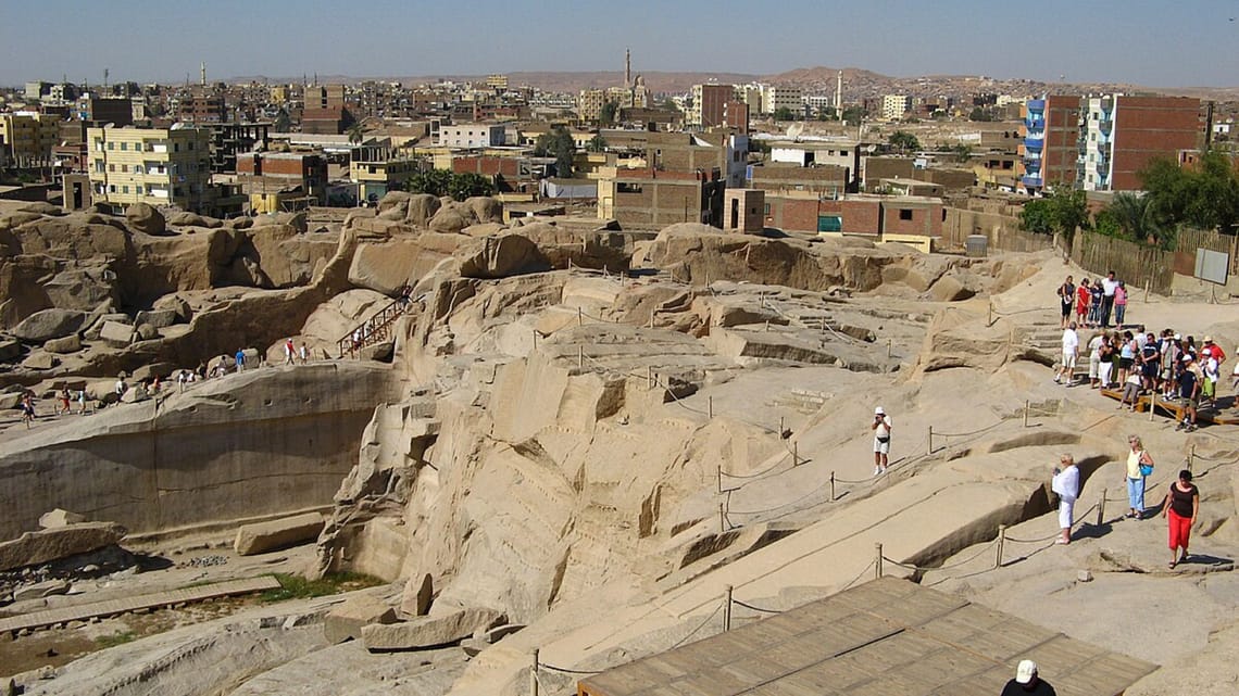 A high-angle view looking down at the massive Unfinished Obelisk carved directly into the pink granite bedrock of an ancient quarry in Aswan, under a clear sky. Caption A fascinating glimpse into ancient craftsmanship: the monumental Unfinished Obelisk