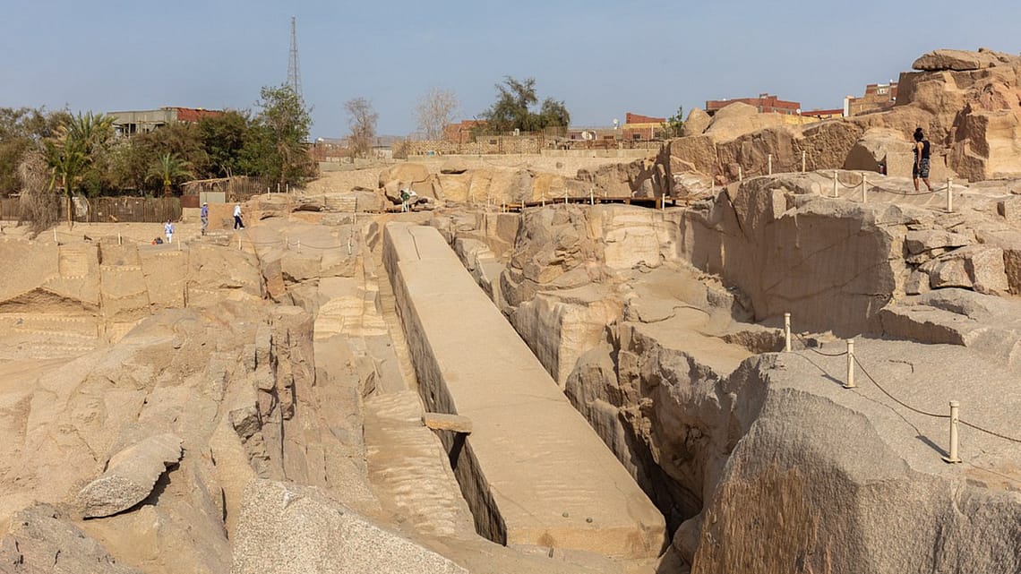 A high-angle view looking down at the massive Unfinished Obelisk carved directly into the pink granite bedrock of an ancient quarry in Aswan, under a clear sky. Caption A fascinating glimpse into ancient craftsmanship: the monumental Unfinished Obelisk