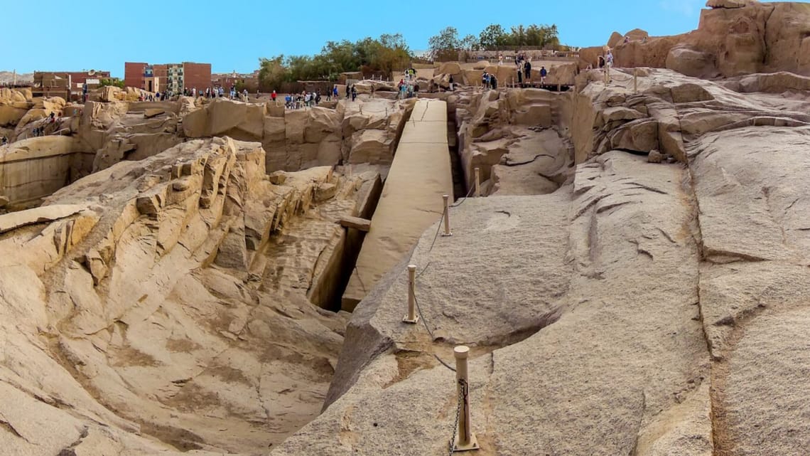 A high-angle view looking down at the massive Unfinished Obelisk carved directly into the pink granite bedrock of an ancient quarry in Aswan, under a clear sky. Caption A fascinating glimpse into ancient craftsmanship: the monumental Unfinished Obelisk