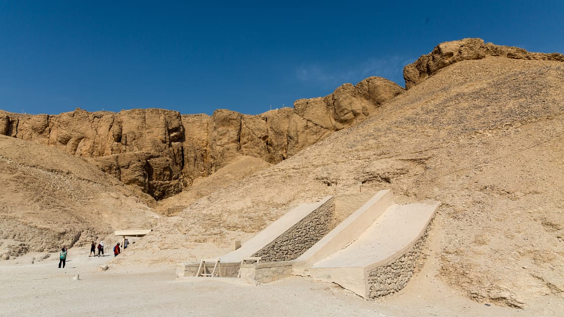 A wide daytime view of the desert landscape at the Valley of the Kings in Luxor, showing two concrete-shaded ramps leading down into underground ancient tombs carved into the limestone hills.