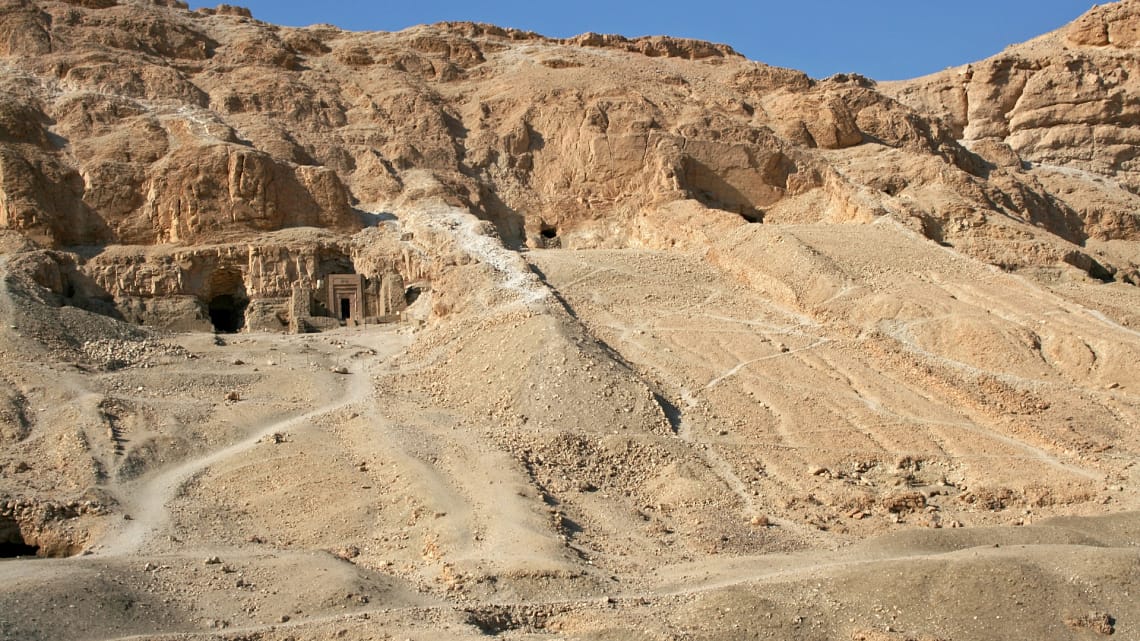 A wide daytime shot focusing on the rugged, sandy limestone cliffs of the Valley of the Kings in Luxor, showing a dirt path leading up to ancient rock-cut tomb openings on the hillside.