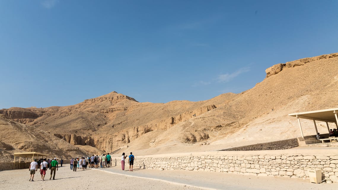 A wide daytime view of the arid, rocky desert hills of the Valley of the Kings in Luxor, with a group of tourists walking along a paved visitor pathway.