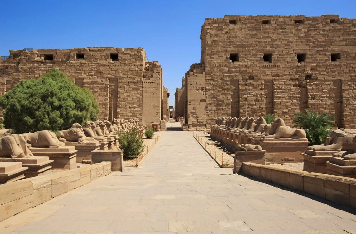 The stone pathway leading to the massive pylon entrance of Karnak Temple in Luxor, lined on both sides by ancient ram-headed sphinx statues under a bright blue sky.