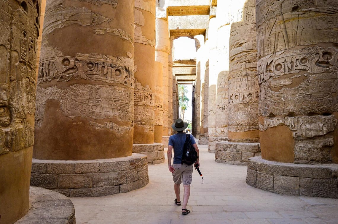 A Top Class Egypt Travel client, wearing a hat and carrying gear, walks between the massive, hieroglyph-carved columns of the Great Hypostyle Hall at Karnak Temple during a private, expert-led tour in Luxor