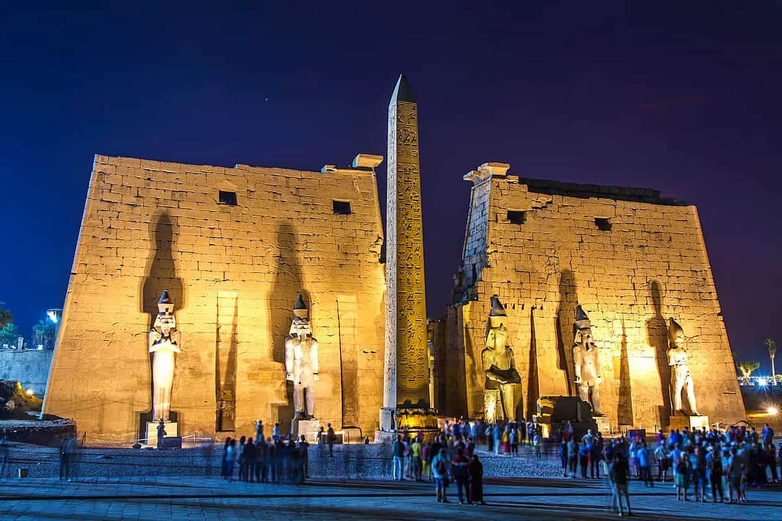 A wide-angle night view of the brilliantly illuminated First Pylon of Luxor Temple, featuring towering seated statues of Ramesses II and a dramatic stone obelisk under a dark evening sky.