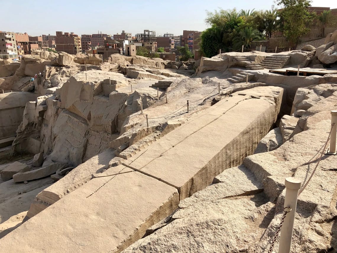 A high-angle view looking down at the massive Unfinished Obelisk carved directly into the pink granite bedrock of an ancient quarry in Aswan, under a clear sky. Caption A fascinating glimpse into ancient craftsmanship: the monumental Unfinished Obelisk