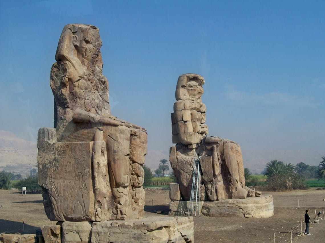 A daytime shot of the two massive weathered stone statues known as the Colossi of Memnon, seated majestically on the West Bank of Luxor against a clear blue sky.