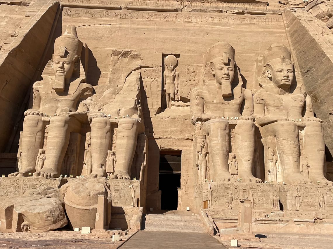 A front-facing daytime view of the Great Temple of Abu Simbel, showcasing the towering carved stone colossi statues of Pharaoh Ramesses II guarding the rock-cut entrance.