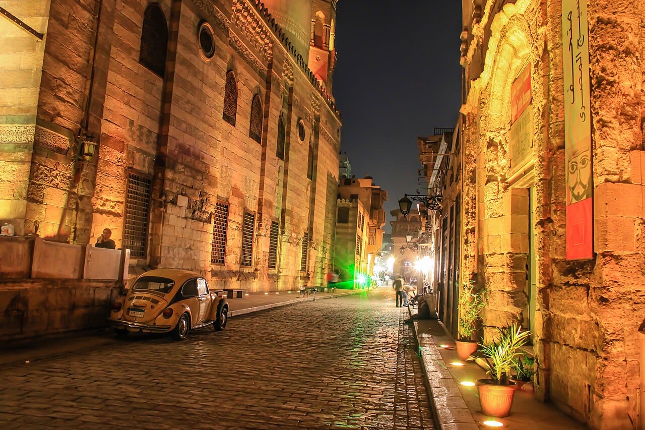 A charming historic cobblestone street in Islamic Cairo at night, featuring illuminated ancient stone madrasas, a vintage yellow Volkswagen Beetle parked on the side, and pedestrian activity under a warm golden evening glow.