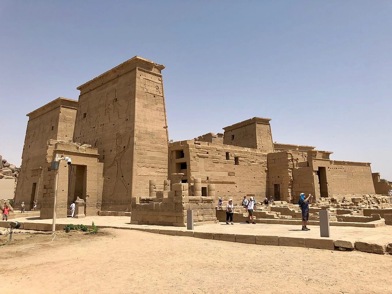 A wide daytime view of the stone pylons and ancient courtyards of the Temple of Isis on Agilkia Island in Aswan, with visitors walking near the historic ruins under a clear sky.