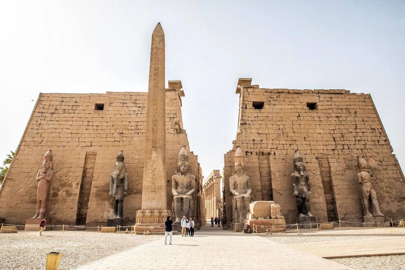 The grand entrance pylon of Luxor Temple with ancient stone obelisks and statues under a bright sky.