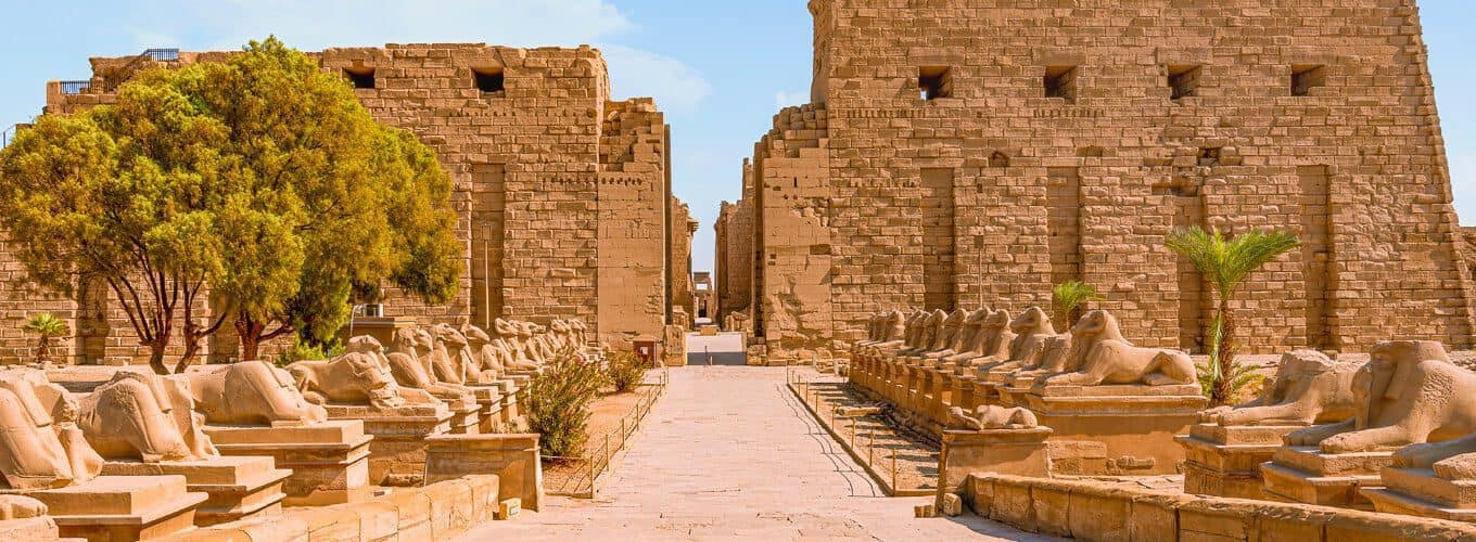 The grand entrance pylon of Karnak Temple in Luxor, featuring the Avenue of Ram-headed Sphinxes under a clear blue sky.