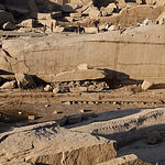 A high-angle view looking down at the massive Unfinished Obelisk carved directly into the pink granite bedrock of an ancient quarry in Aswan, under a clear sky. Caption A fascinating glimpse into ancient craftsmanship: the monumental Unfinished Obelisk