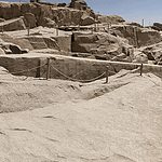 A high-angle view looking down at the massive Unfinished Obelisk carved directly into the pink granite bedrock of an ancient quarry in Aswan, under a clear sky. Caption A fascinating glimpse into ancient craftsmanship: the monumental Unfinished Obelisk