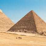 A wide panoramic view of the Great Pyramids of Giza and the Queens' Pyramids with camels in the desert, part of a private luxury itinerary by Top Class Egypt Travel.