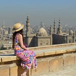 A woman in a sun hat sitting on a stone wall overlooking the domes and minarets of Islamic Cairo. - Saladin Citadel of Cairo