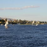 Traditional Egyptian felucca sailboats sailing on the Nile River in Luxor with lush riverbanks and desert hills in the background.9 Days Cairo and Nile cruise.
