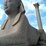 The ancient Roman Pompey's Pillar alongside a granite Sphinx statue under a blue sky in Alexandria Egypt.