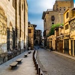 A daytime perspective of the historic cobblestone Al-Muizz Street in Old Cairo, lined with monumental medieval Islamic stone walls, traditional wooden windows, and local shopfronts under a soft sky.