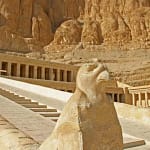A close-up shot of a weathered stone Horus falcon statue standing guard at the base of a grand stone ramp, leading up to the tiered terraces and colonnades of the Mortuary Temple of Hatshepsut against the steep limestone cliffs of Deir el-Bahari.
