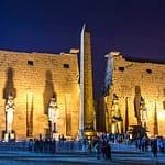 A wide-angle night view of the brilliantly illuminated First Pylon of Luxor Temple, featuring towering seated statues of Ramesses II and a dramatic stone obelisk under a dark evening sky.