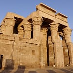 A warm, low-angle daytime view of the ancient stone portal and heavy carved columns of the Temple of Kom Ombo against a clear sky.