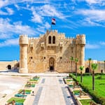 A wide daytime view of the prominent stone fortress of the Citadel of Qaitbay in Alexandria, featuring its fortified towers, main entrance, and a landscaped green garden under a bright blue sky with light clouds.