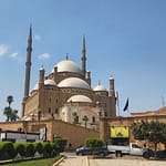 A low-angle view looking up at the monumental Muhammad Ali Mosque within the Citadel of Salah El-Din in Cairo, showcasing its grand stone domes, twin pencil minarets, and historic fortified walls against a blue sky.