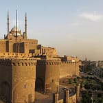 Panoramic view of the historic stone walls of the Citadel of Salah El-Din in Cairo, featuring the prominent domes and minarets of the Mohamed Ali Mosque against a clear sky.