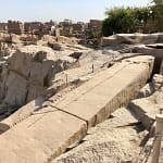 A high-angle view looking down at the massive Unfinished Obelisk carved directly into the pink granite bedrock of an ancient quarry in Aswan, under a clear sky. Caption A fascinating glimpse into ancient craftsmanship: the monumental Unfinished Obelisk