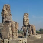 A daytime shot of the two massive weathered stone statues known as the Colossi of Memnon, seated majestically on the West Bank of Luxor against a clear blue sky.