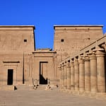 A wide, front-facing view of the massive stone pylon gateway at the Philae Temple of Isis in Aswan, with a row of beautifully carved stone columns lining the courtyard to the right under a bright blue sky.