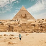 A person walking towards the Great Sphinx of Giza with the Pyramid of Khafre in the background