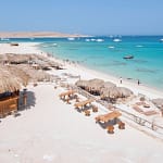 A high-angle daytime view of a white sand beach resort in Hurghada, featuring thatched-roof umbrellas, sun loungers, and clear turquoise Red Sea water with boats anchored in the distance.