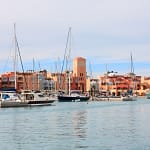 A scenic daytime view of El Gouna Marina featuring luxury yachts and sailboats moored in the calm harbor waters, with Mediterranean-style terracotta and pastel resort buildings lining the waterfront.