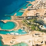 A high-angle aerial view of the Red Sea coastline in Hurghada, showcasing deep blue waters shifting to a turquoise lagoon beach and a premium resort hotel layout along the coast.